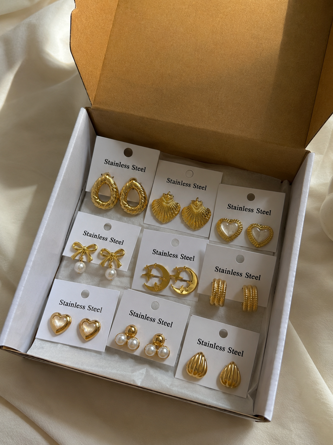 A boxed set of gold and pearl stud earrings displayed on white cards labeled 'Stainless Steel' inside an open brown gift box on a light fabric surface.